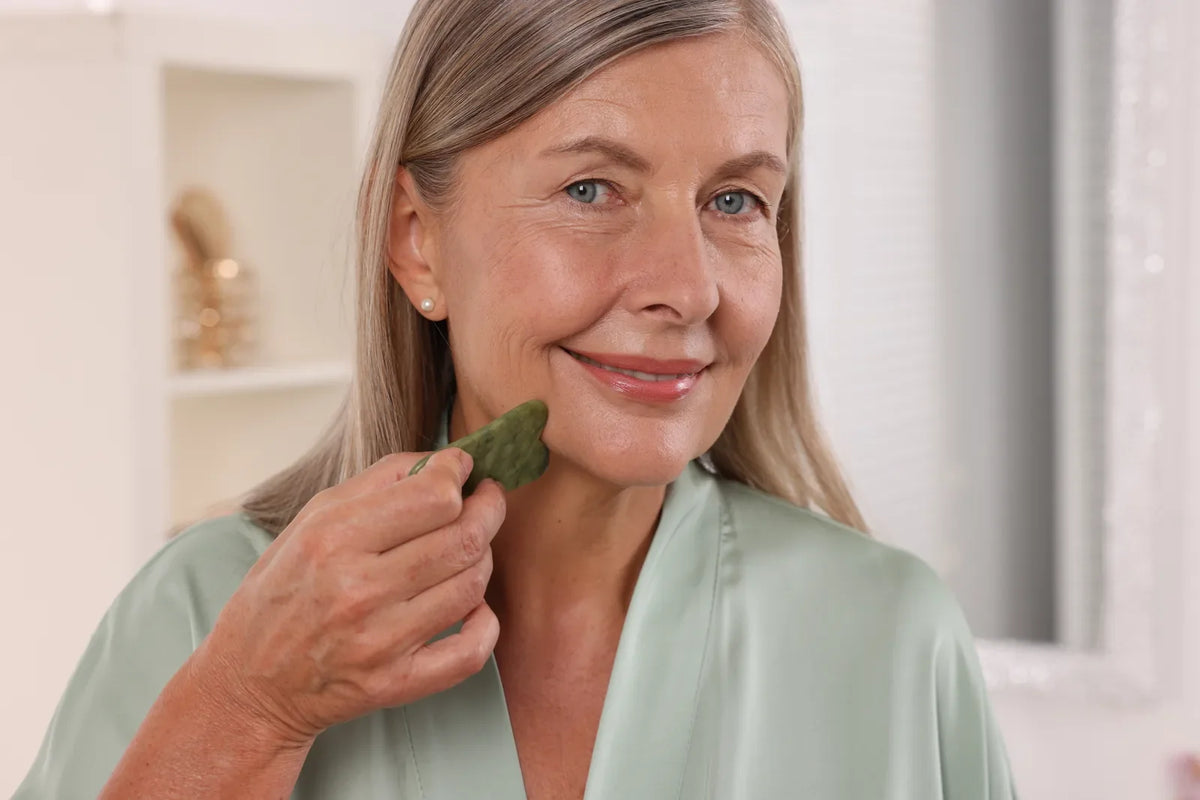 Woman with silver hair gliding a green jade gua sha stone along her jaw toward her ear in a bright bathroom