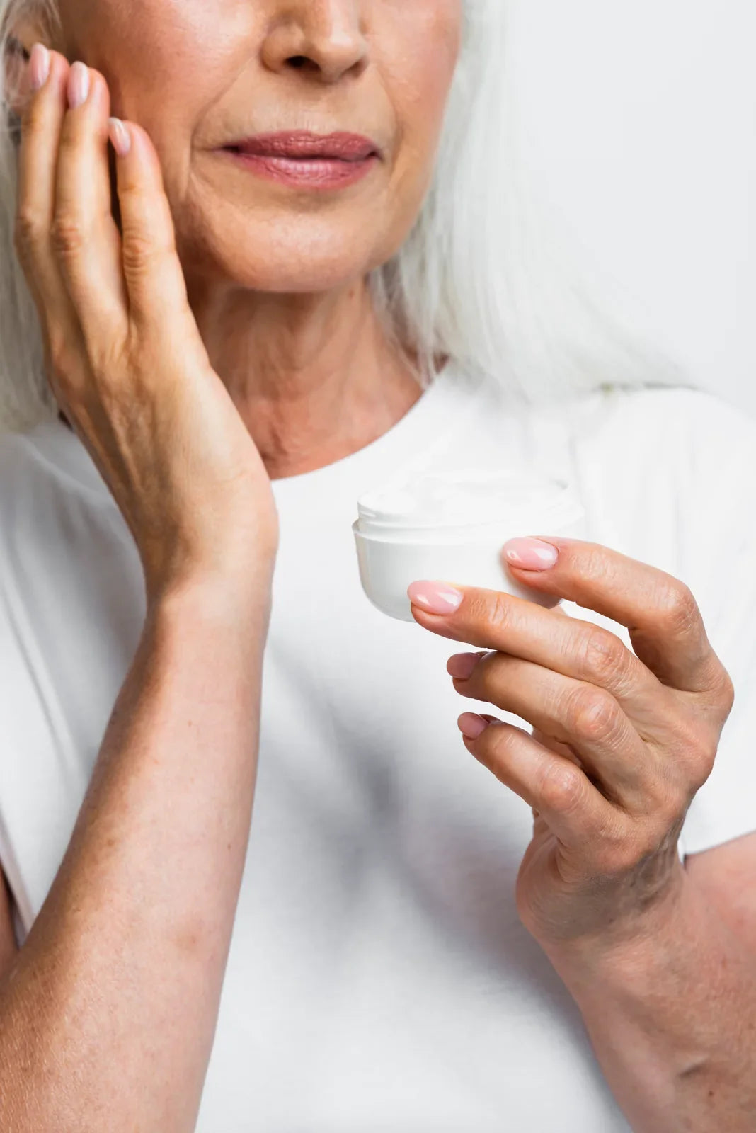 Woman with silver hair pressing a rich face cream into her cheek, holding an open unbranded jar
