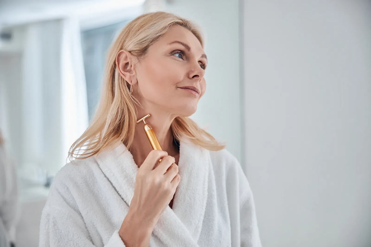 Woman in her fifties gliding a slim handheld facial massager along her jawline in a bright bathroom