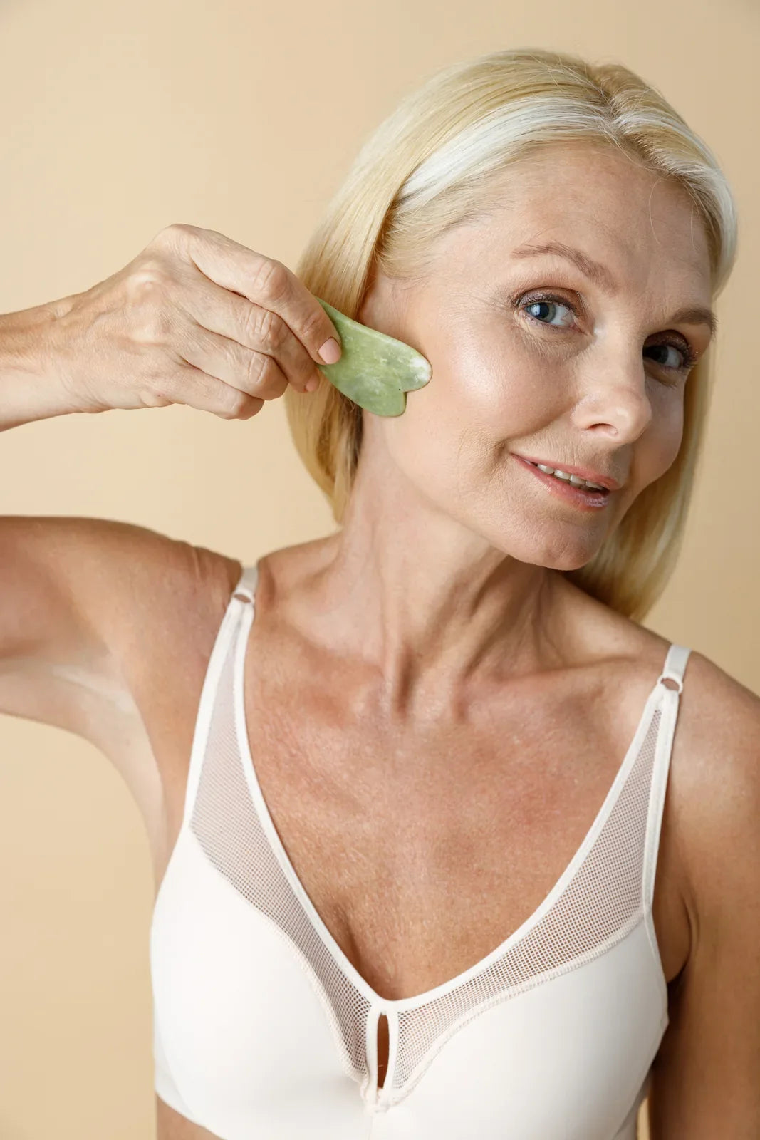 Woman in her fifties gliding a green jade gua sha stone along her cheekbone