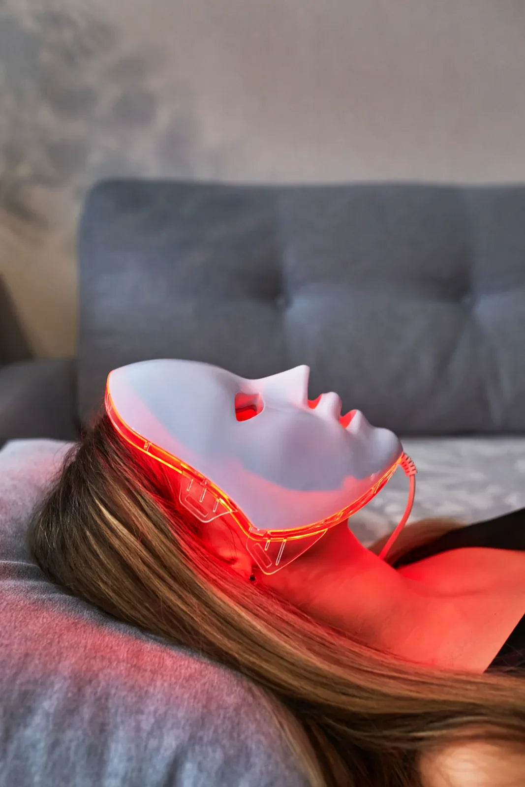 Woman relaxing on a sofa at home wearing an LED face mask glowing with red light